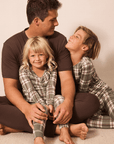 A father and two children wearing matching brown and green plaid pajamas sitting together on a light beige floor against a plain beige wall.