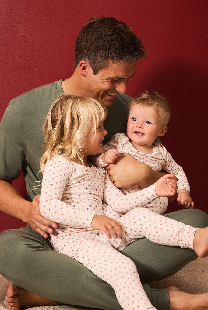 A father and two children wearing matching light red and white twinkle star pajamas, sitting together against a solid dark red background.