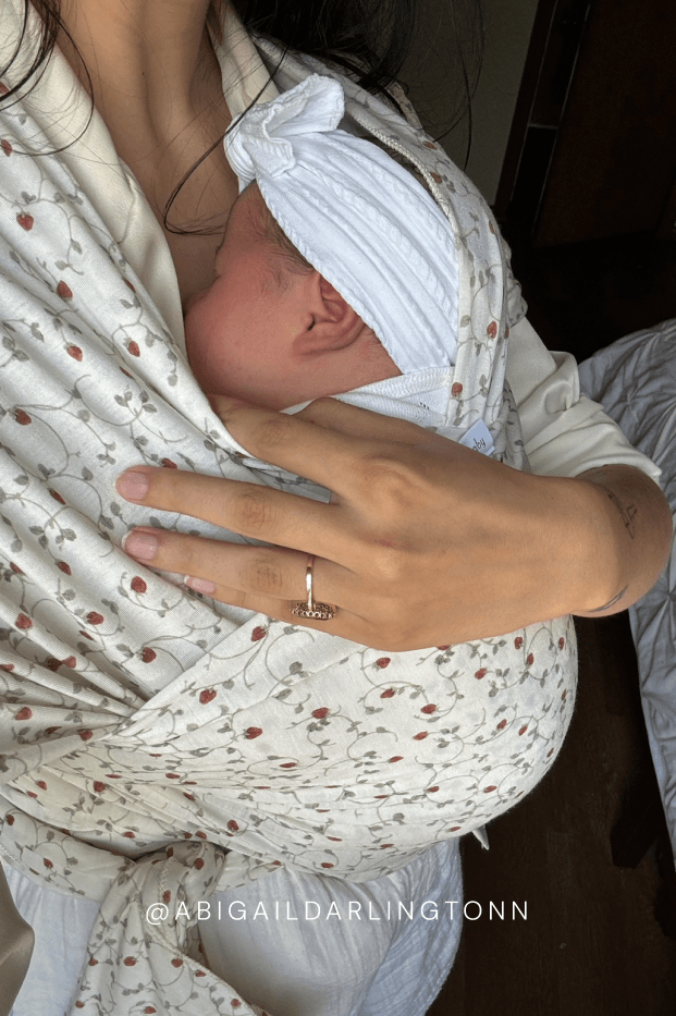 Newborn baby in a strawberry baby carrier with a person holding it, wearing a ring. @abigaildarlingtonn