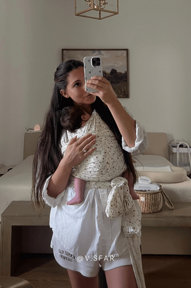 Woman holding a baby in a strawberry wrap and taking a mirror selfie in a home setting. @v.sfar