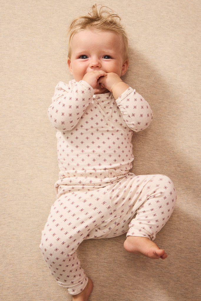 A baby lying on a beige textured blanket, wearing a cream-colored long-sleeve romper and matching pants with a small red star pattern.