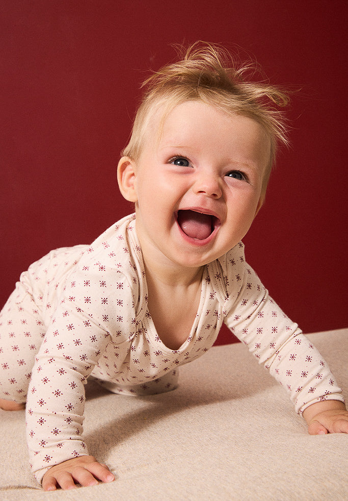 A baby crawling on a beige fabric surface against a solid dark red background, wearing a cream-colored long-sleeve bodysuit with a small red star pattern.