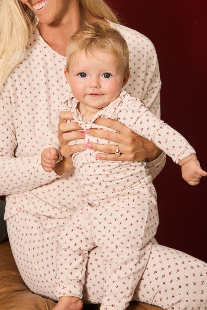 A baby and mother wearing matching long-sleeve pajamas with a small red star pattern on a cream background, posed against a dark red background.
