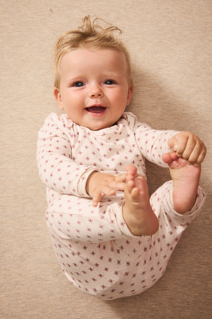 A smiling baby lying on a beige textured blanket, wearing a light red long-sleeve onesie with a small red star pattern.