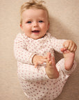 A smiling baby lying on a beige textured blanket, wearing a light red long-sleeve onesie with a small red star pattern.