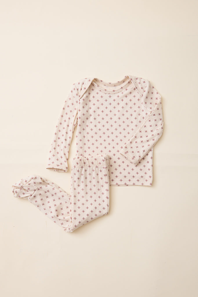 A two-piece baby pajama set featuring a long-sleeve top and matching footed pants, both in a light cream fabric with a small red star pattern, laid flat on a warm beige background.