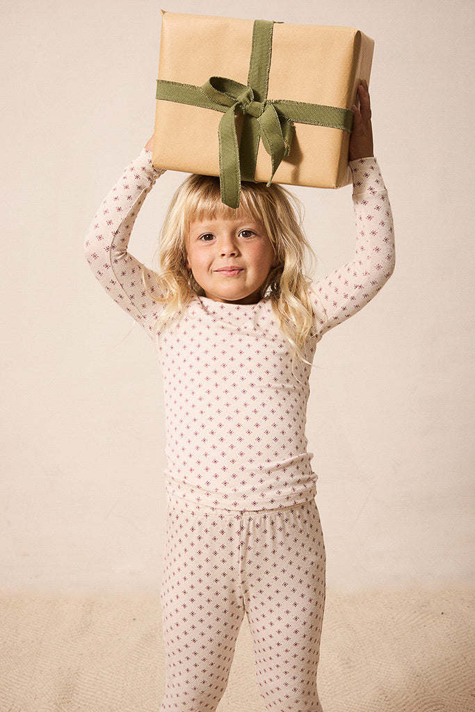 A young blonde girl wearing a cream-colored long-sleeve pajama set with small red star pattern holds a wrapped gift box with a green ribbon on her head, posing against a warm beige studio background.