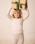 A young blonde girl wearing a cream-colored long-sleeve pajama set with small red star pattern holds a wrapped gift box with a green ribbon on her head, posing against a warm beige studio background.