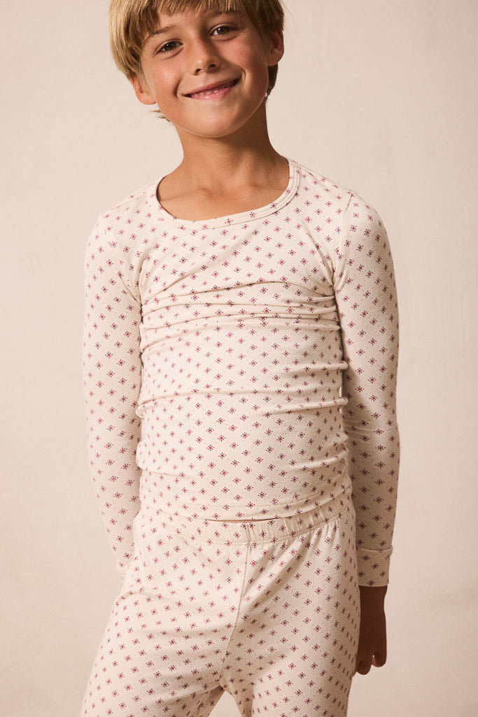 A smiling young boy wearing a long-sleeve pajama top and matching pants set in a cream color with a small red star pattern, set against a plain beige background.