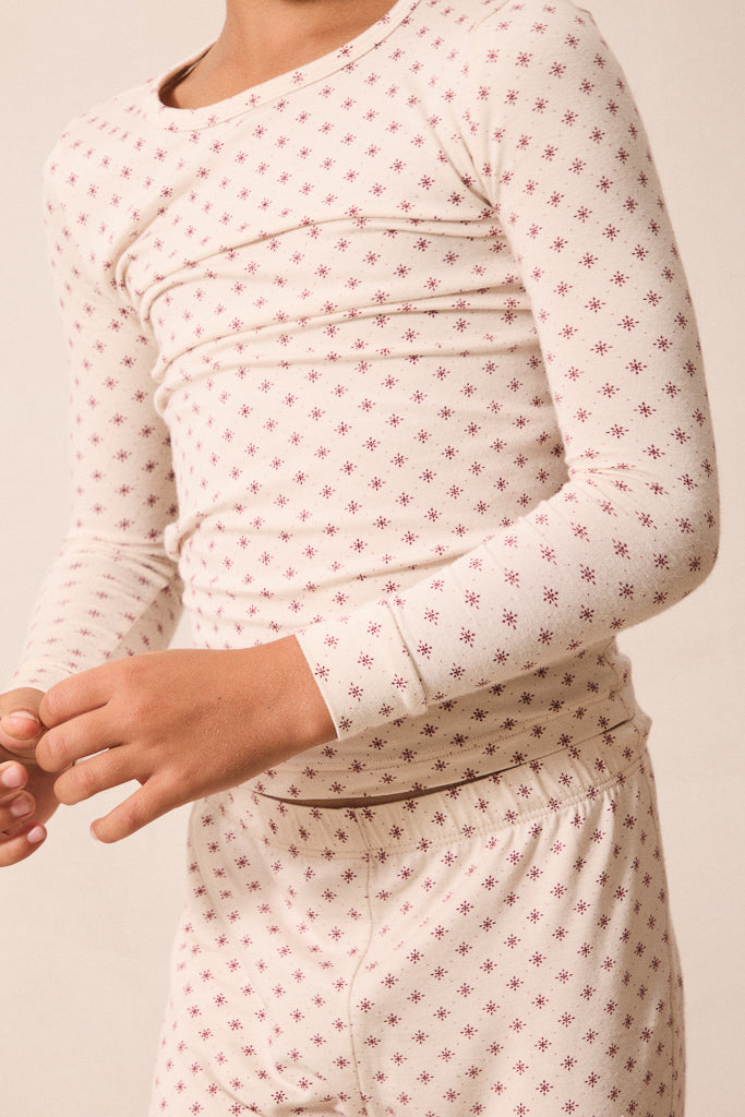 A close-up of a person wearing a long-sleeve cream-colored pajama top with a small repeating red star pattern, paired with matching pajama pants against a plain light beige background.