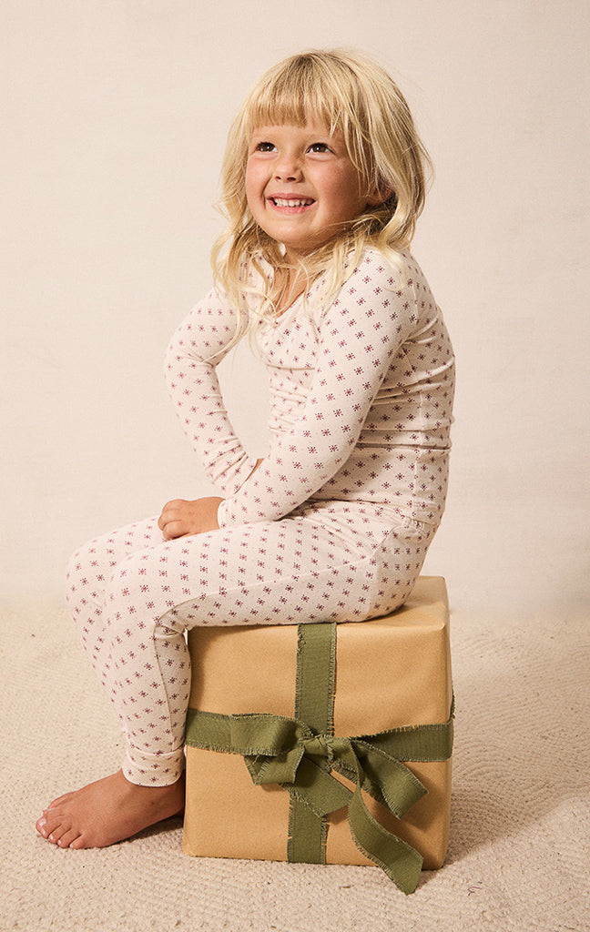 A smiling young girl with blonde hair sits barefoot on a brown paper gift box with an olive green ribbon. She is wearing a cream-colored long-sleeve pajama top and matching pants with a small red star pattern.