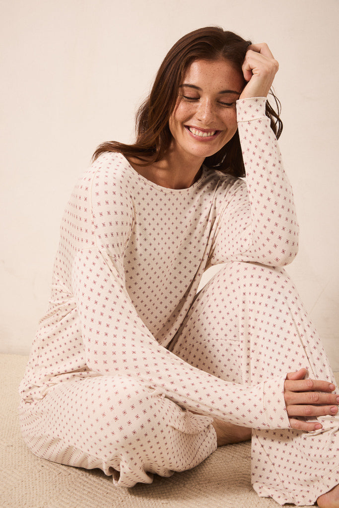 A woman sitting on a textured beige floor wearing a matching long-sleeve pajama set with a small red star pattern on a cream background.