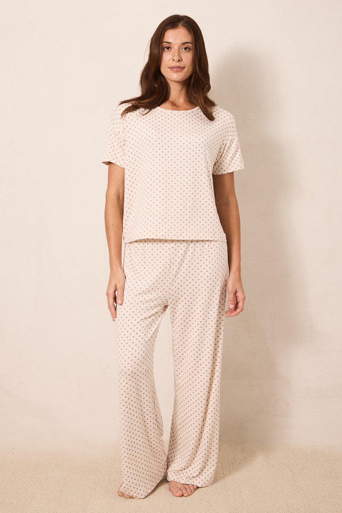 A woman modeling a two-piece pajama set consisting of a short-sleeve top and matching wide-leg pants, both in a light beige color with a small red star pattern, set against a plain beige studio background.