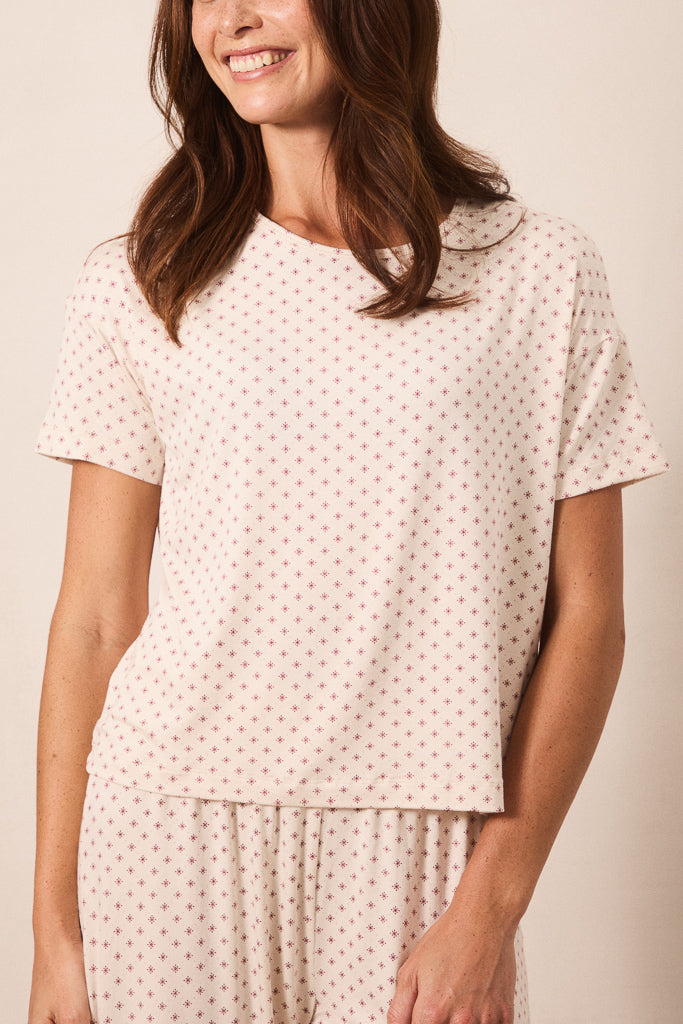 A woman wearing a short-sleeve crewneck pajama top and matching pants in a cream color with a small red star pattern, photographed against a solid beige background.