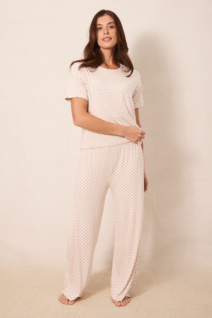 A woman modeling a matching two-piece loungewear set consisting of a short-sleeve top and wide-leg pants, both in a light cream color with a small red star pattern, against a plain beige studio background.