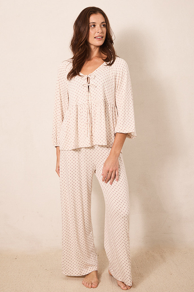 A woman modeling a two-piece pajama set consisting of a long-sleeve peplum top and matching wide-leg pants, both in a cream color with a small red star pattern, against a warm beige studio background.
