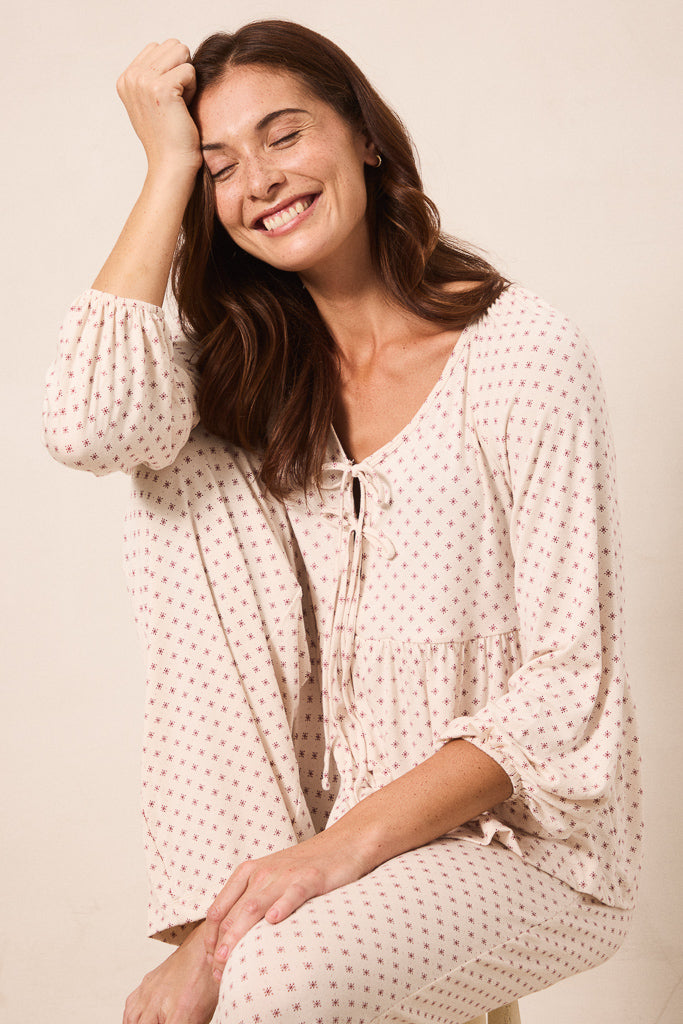 A woman wearing a cream-colored, long-sleeve tie-front top and matching pants with a small red star pattern, posing against a warm beige background.