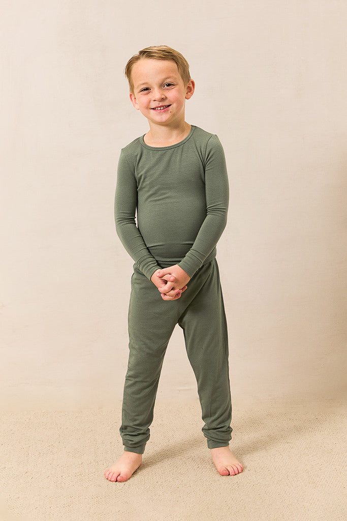 A young boy smiling and standing barefoot, wearing a matching two-piece pajama set in olive green against a warm beige studio backdrop.
