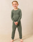 A young boy smiling and standing barefoot, wearing a matching two-piece pajama set in olive green against a warm beige studio backdrop.