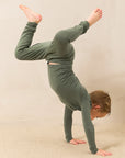 A child wearing an olive green long-sleeve pajama set performs a handstand on a beige carpeted floor against a neutral beige wall.