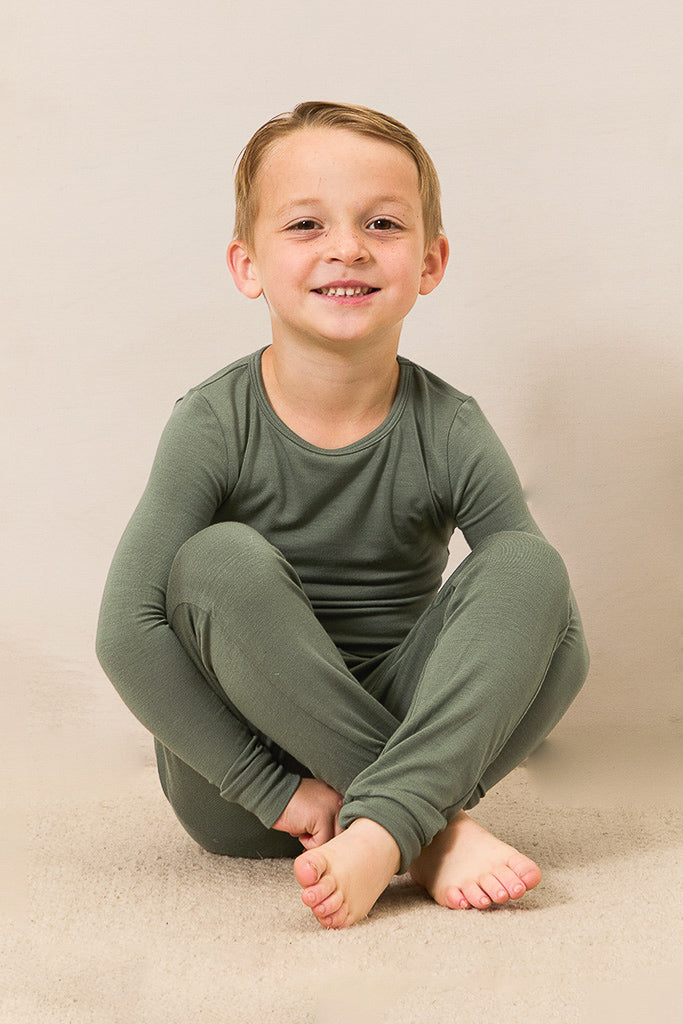 A young boy sitting cross-legged on a beige carpeted floor against a neutral beige background, wearing a matching long-sleeve crewneck pajama set in olive green.