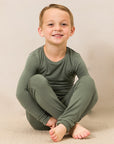 A young boy sitting cross-legged on a beige carpeted floor against a neutral beige background, wearing a matching long-sleeve crewneck pajama set in olive green.