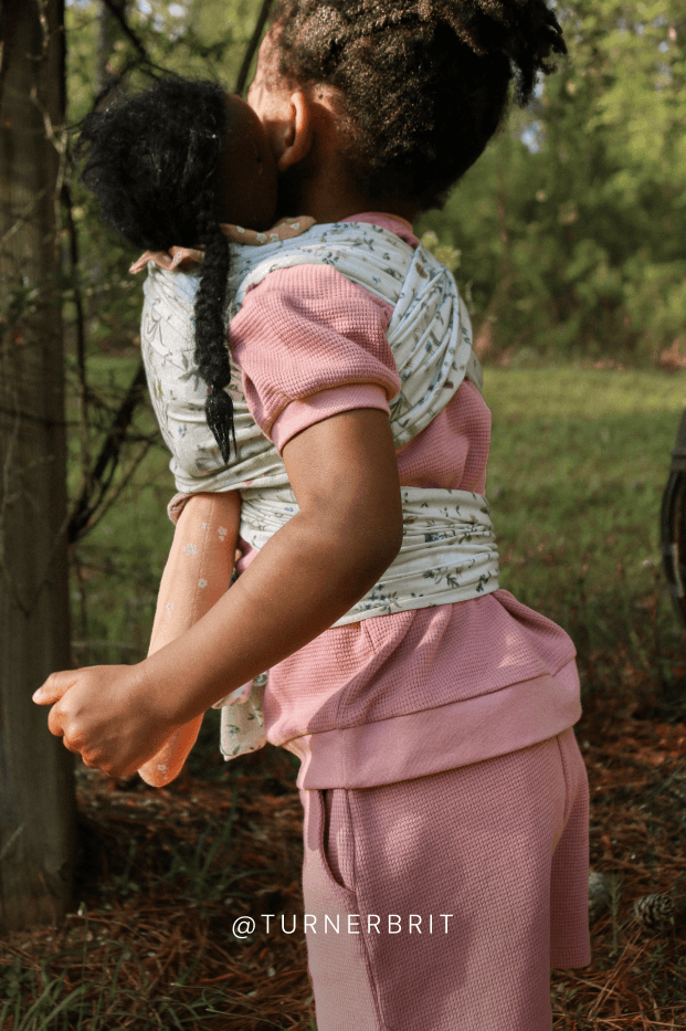 Child carrying her dolly in birthday botanical baby wrap play carrier outdoors. Text reads @TURNERBRIT