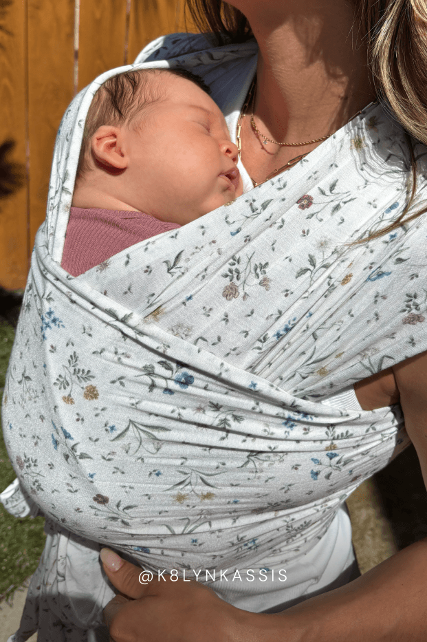 Newborn baby in a floral baby carrier with a blurred background. Text reads @k8LYNKASSIS
