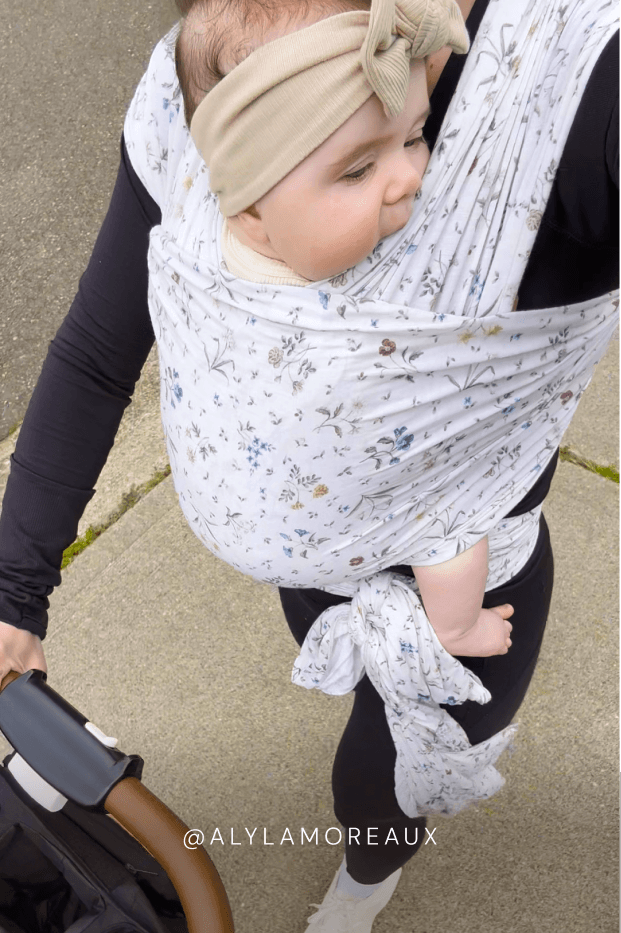 Mom holding a baby in a floral baby carrier on a sidewalk while pushing a stroller. Text reads @ALYLAMOREUX