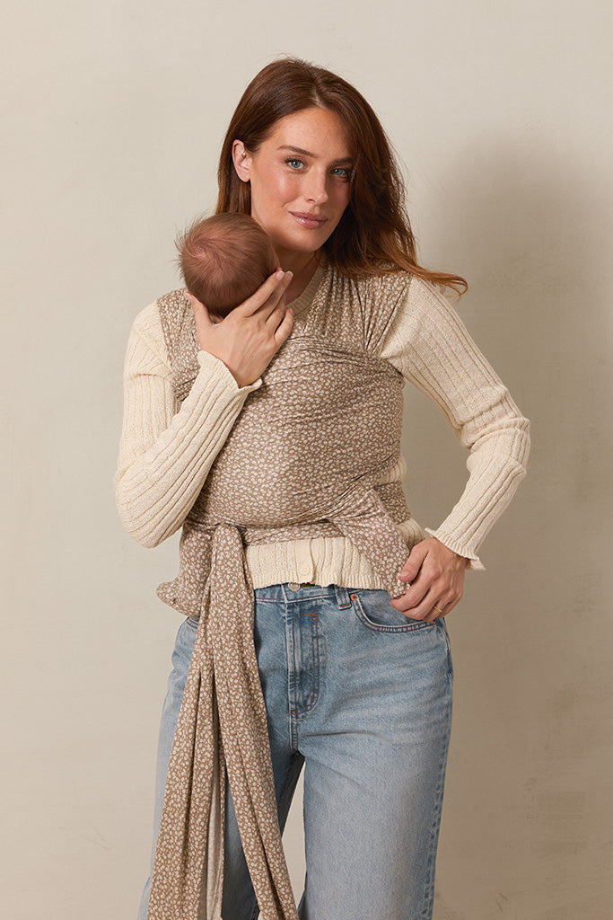 A woman wearing a white blouse and jeans standing and holding a baby in a Solly Baby Wrap carrier in the Cream Floral print, featuring delicate cream flowers on a taupe background. 