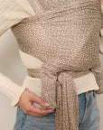 A mother standing while holding her baby in a Solly Baby Wrap carrier in the Cream Floral print, featuring delicate cream flowers on a taupe background. 