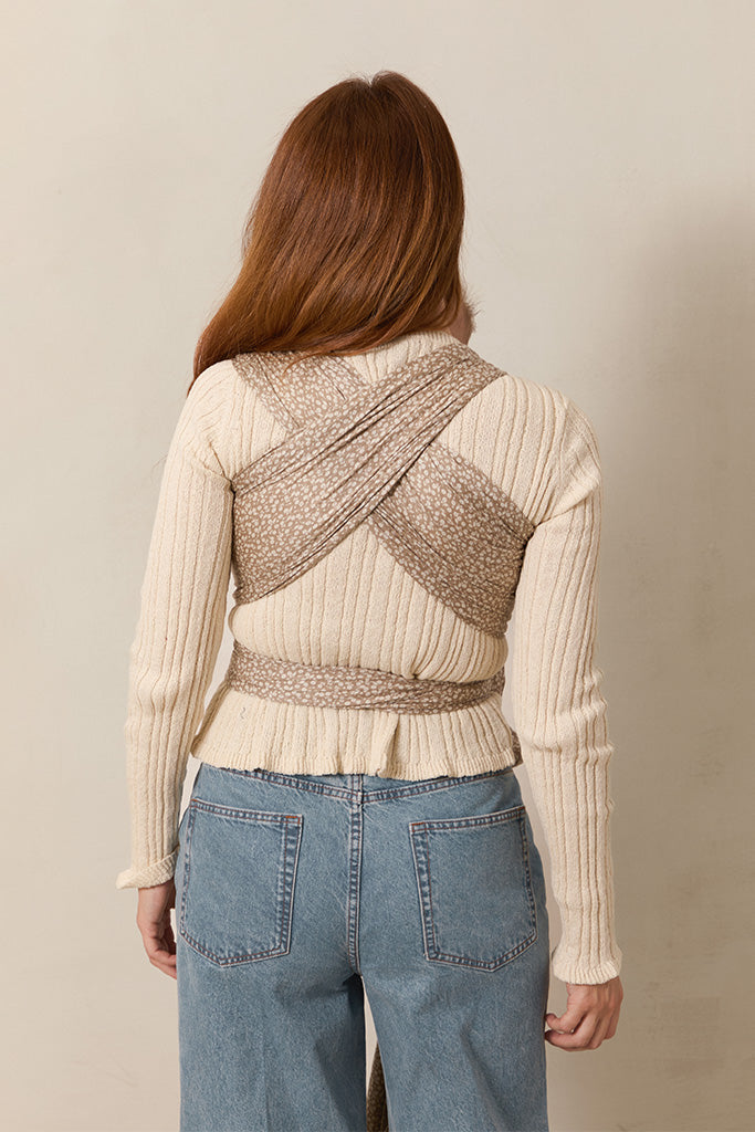 The back view of a woman wearing a Solly Baby Wrap carrier in the Cream Floral print, featuring delicate cream flowers on a taupe background, tied securely at her waist over a cream sweater and jeans.