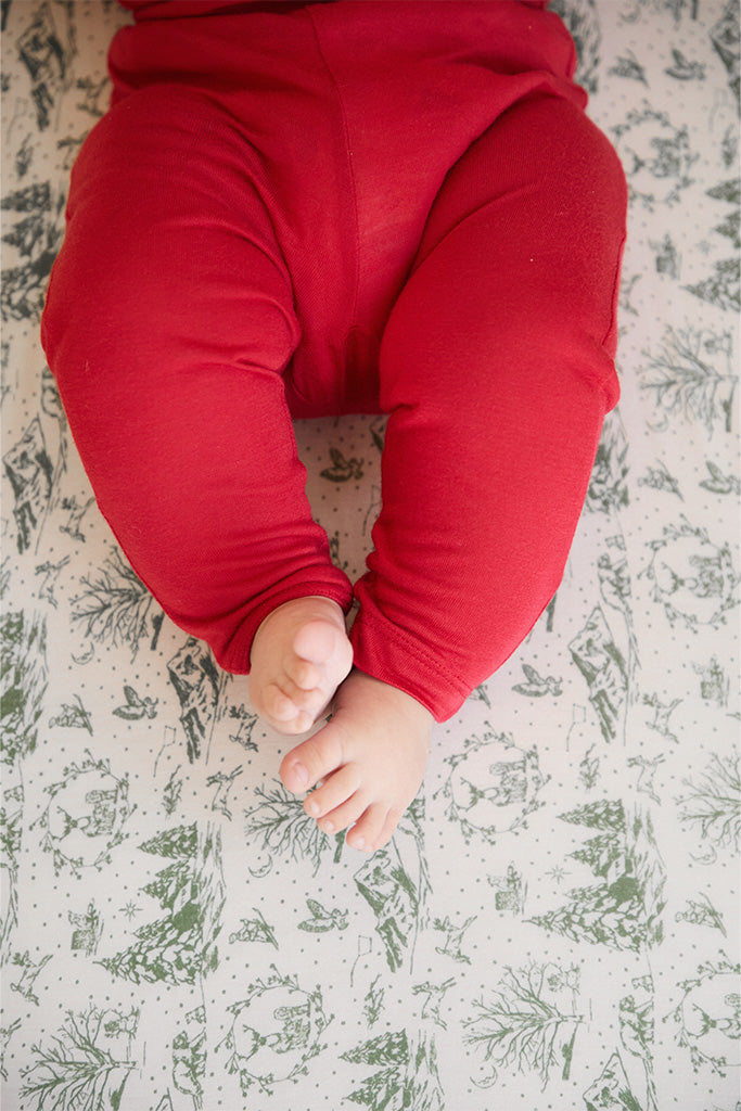 A close-up of the baby pajama bottoms in the Currant color, a vibrant red, resting on a crib sheet featuring a cream and soft green winter forest toile design.