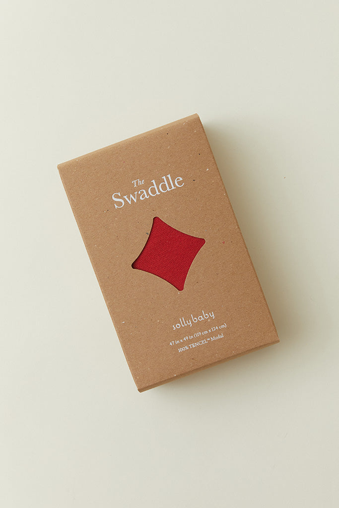 A product packaging shot of the Solly Baby Swaddle in a box with a window showing the Currant colorway inside. 