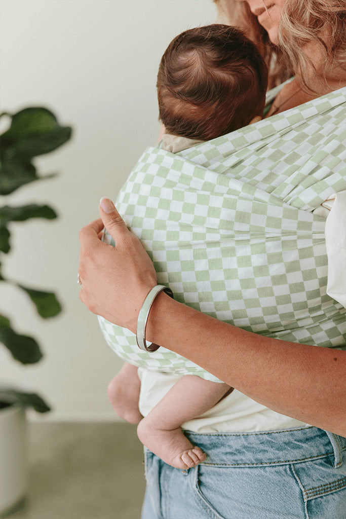 A close-up image of a mother holding her baby in a Solly Baby Wrap carrier in the Fern Chequer print, a soft green and cream checkered print. 