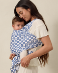 Woman holding a baby wrapped in a blue and white checkered solly baby wrap against a beige background
