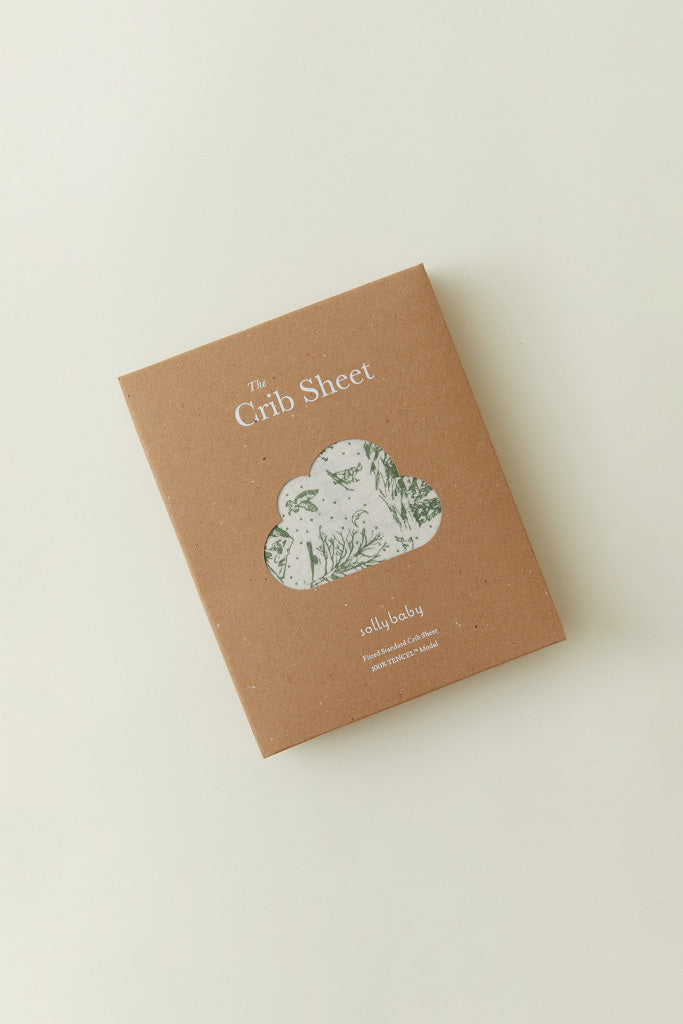 A product packaging shot of the Solly Baby Crib Sheet in a brown box with a window showing the Winter Toile print inside. 