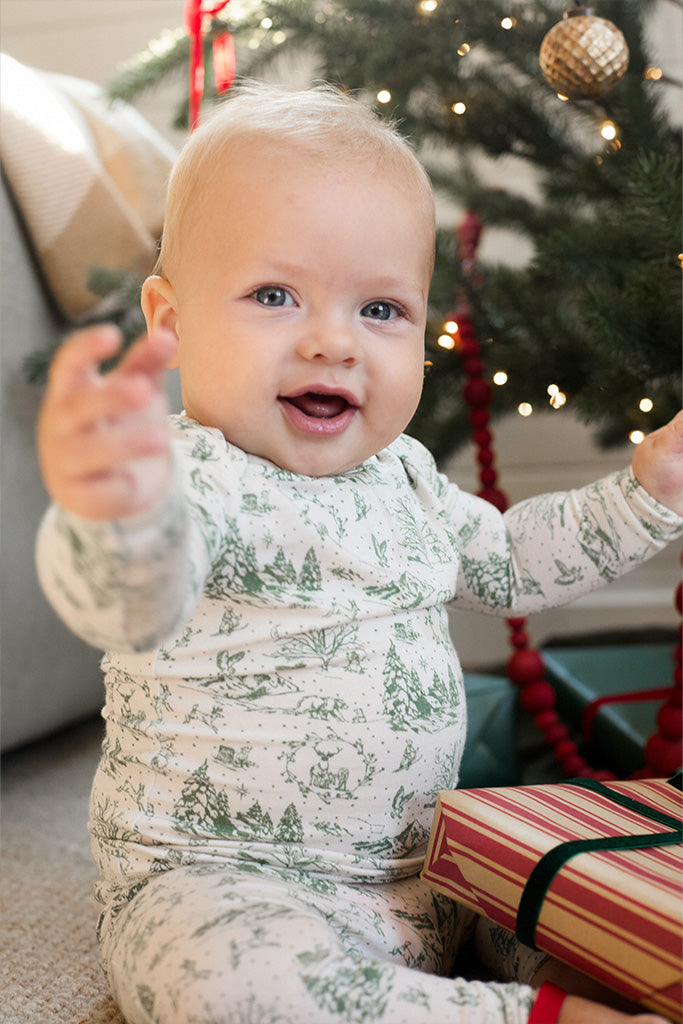 A baby sitting and smiling in front of a Christmas tree, while wearing a matching two-piece pajama set, featuring a cream and soft green winter forest toile design. Sizes range from 0m to 5T.