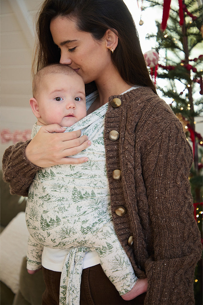 A mother standing while holding her baby in a soft wrap carrier featuring a cream and green winter forest toile design with a cozy Christmas tree in the background.