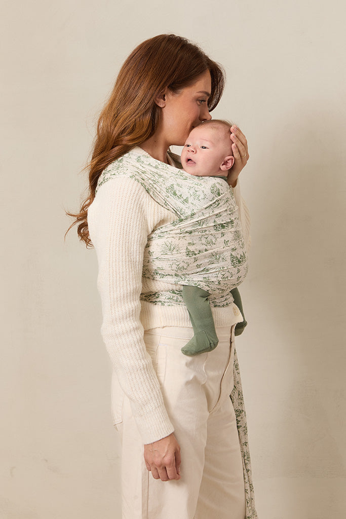 A side view of a woman standing and holding her baby in a soft wrap carrier featuring a cream and green winter forest toile design.